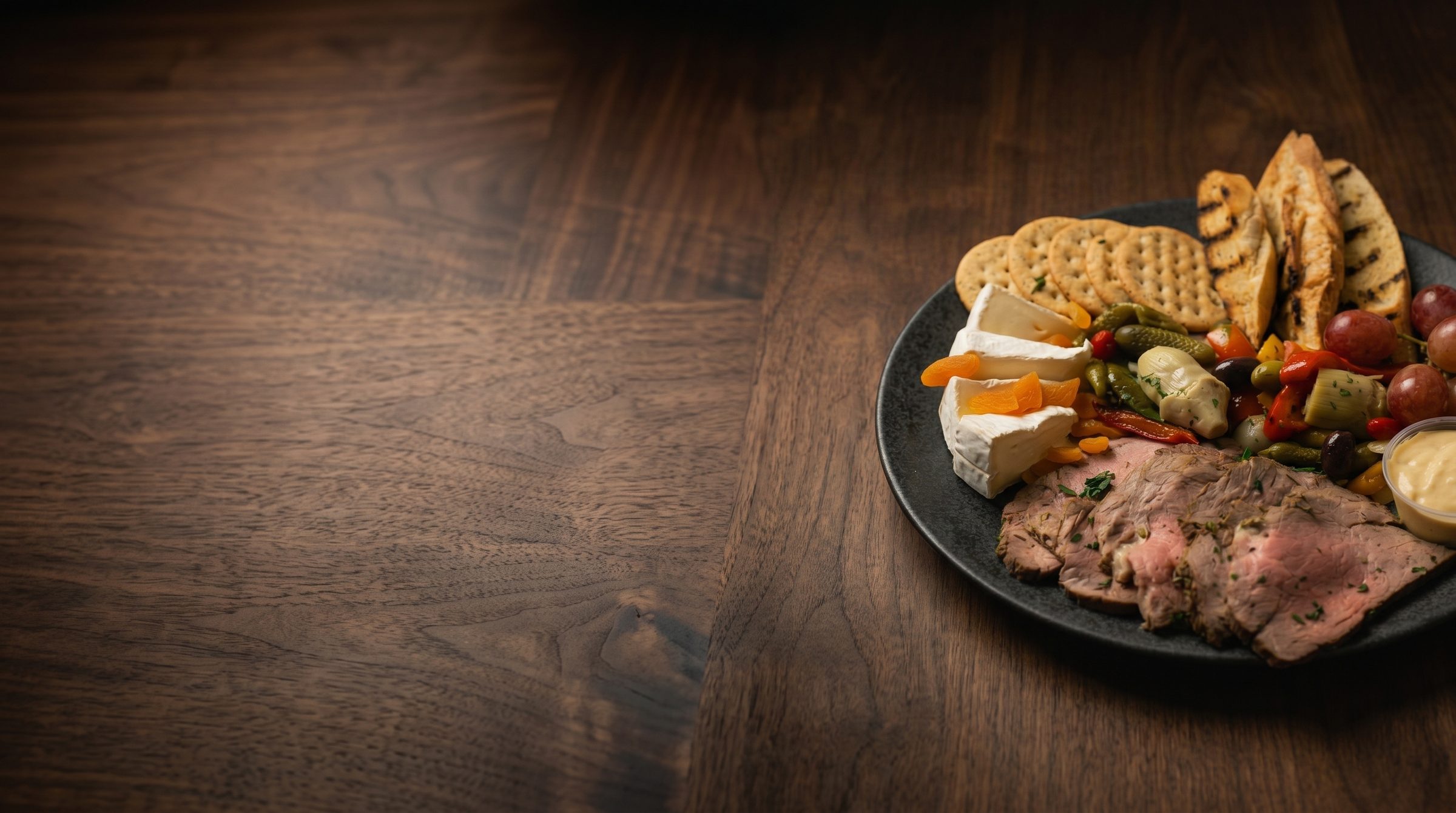 Charcuterie board with sliced roast beef, cheeses, fruit and crackers on dark walnut